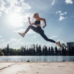 Woman performs an athletic jump by a lakeside in the park under a sunny blue sky.